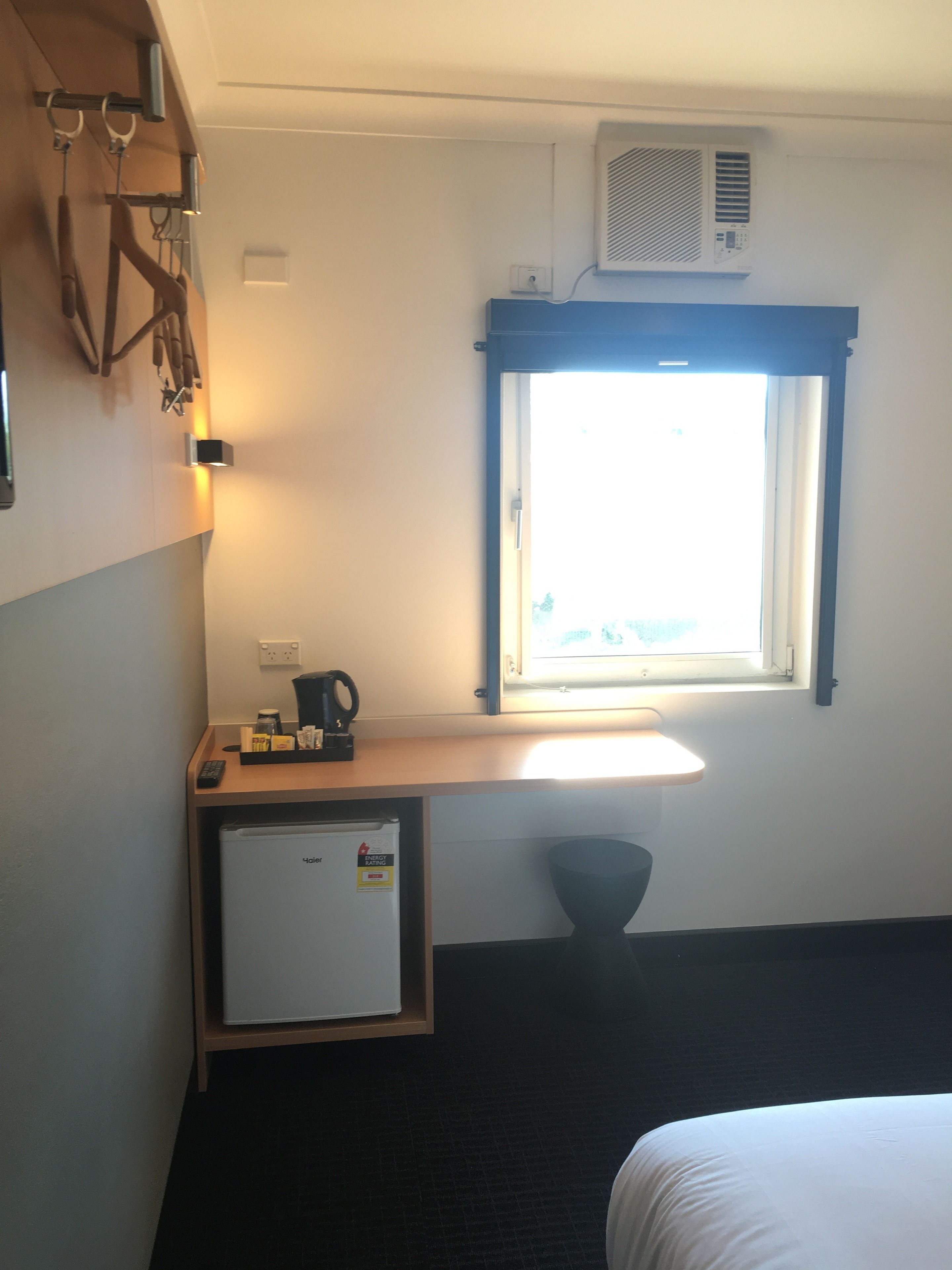 Habitación de hotel compacta con cama, un escritorio pequeño con cafetera, refrigerador minibar, ventana con persianas y unidad de aire acondicionado.