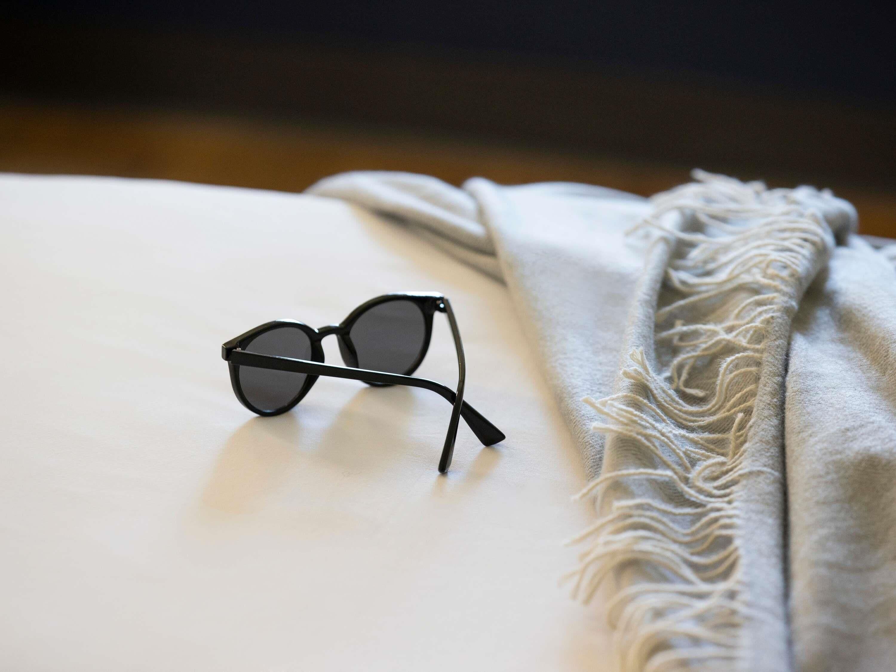 Un par de gafas de montura negra sobre una superficie blanca, con una manta gris texturizada a su lado.