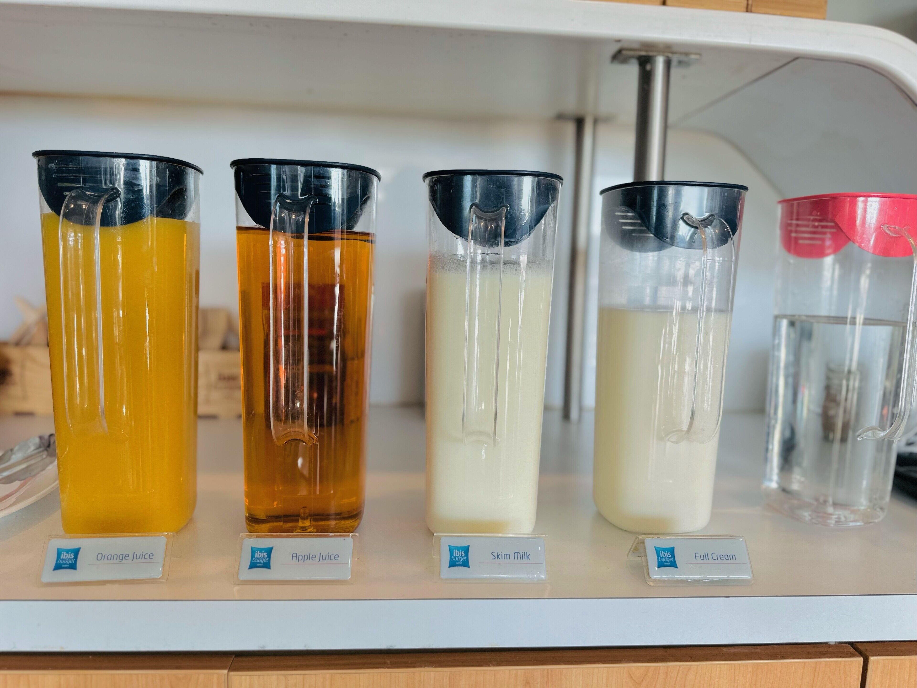 A shelf with four containers labeled with different beverages: orange juice, apple juice, skim milk, and full cream.