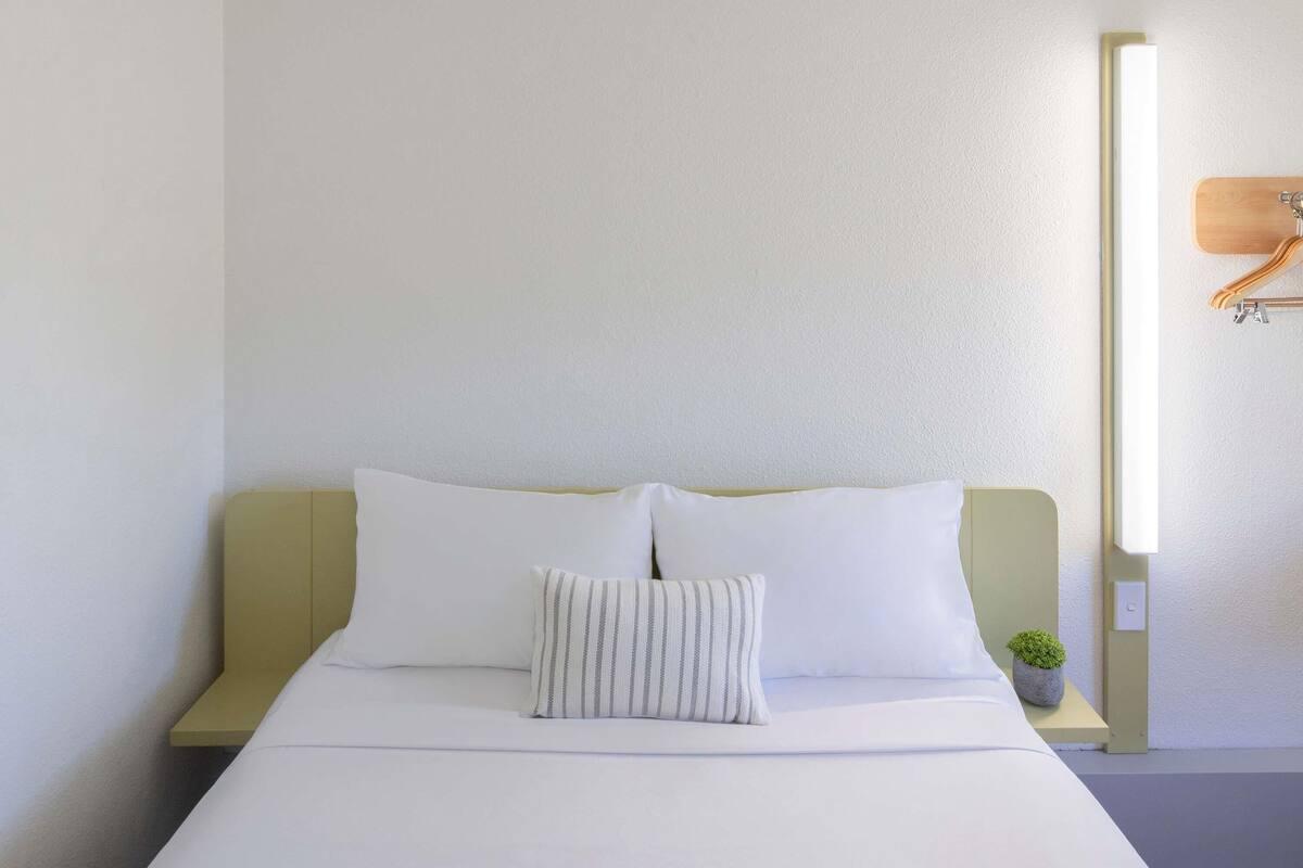 Quarto de hotel minimalista com uma cama, duas almofadas, uma mesa de cabeceira com abajur e uma pequena planta.