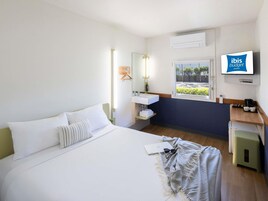Habitación de hotel con cama, mesita de noche con lámpara, ventana con vistas, lavamanos y pantalla de televisión mostrando