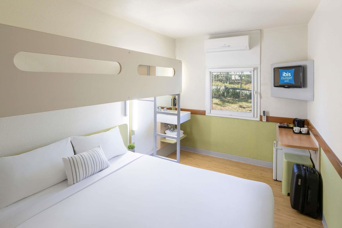 A hotel room with a bed, a small desk with a television, a window, and a trash bin.