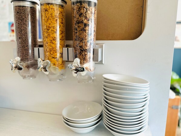 Una estación de servicio de alimentos con tres recipientes de cereales diferentes, una pila de platos blancos y un dispensador de metal.