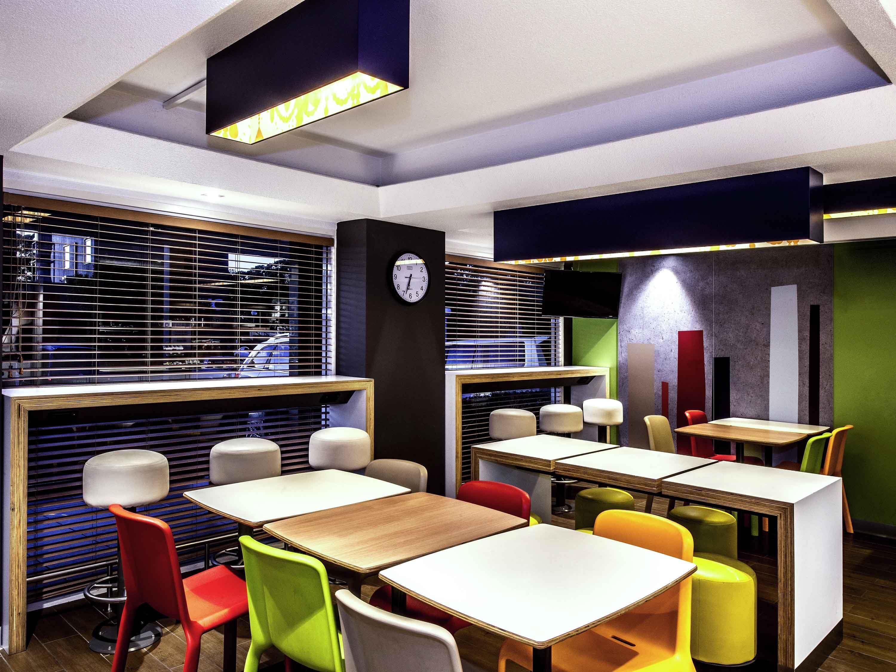 A modern restaurant interior with multiple tables and chairs, a clock on the wall, and a window with blinds.
