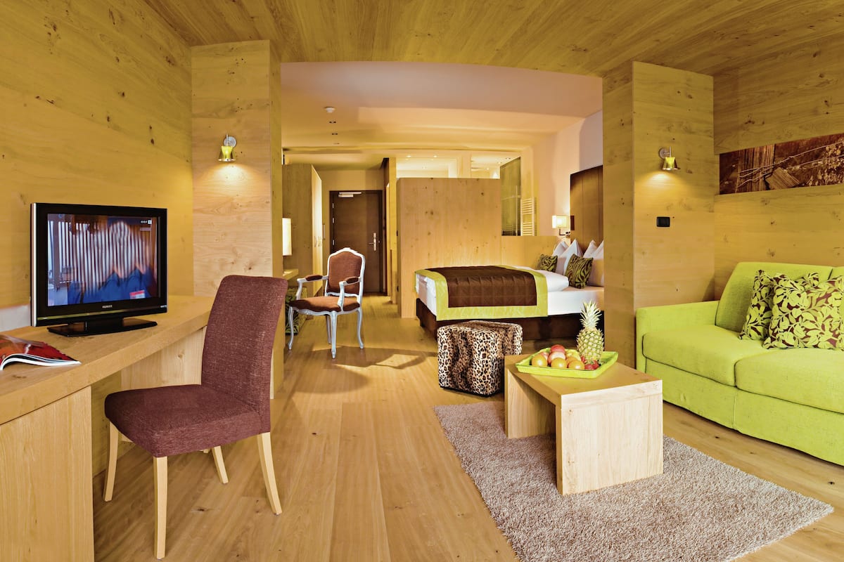Ein modernes Hotelzimmer mit Holzwänden, einem grünen Sofa, einem Holztisch, einem Bestuhl mit Leopardenmuster, einem Fernseher und einer Essecke mit Stuhl und Tisch.