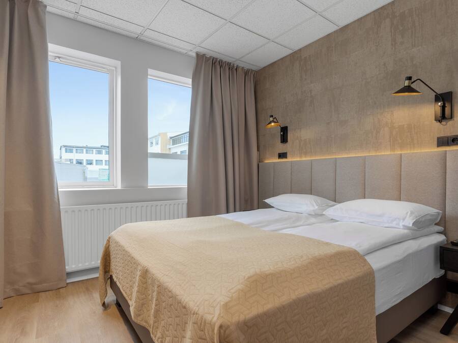 Una habitación de hotel moderna con una cama grande, una mesita de noche, una lámpara de pared y una ventana con cortinas.