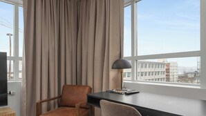 Una habitación con un escritorio, dos sillas, una lámpara y vistas a edificios a través de amplias ventanas.