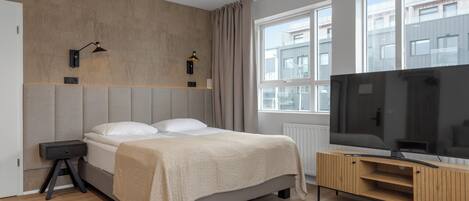 Une chambre d’hôtel moderne avec un grand lit, un téléviseur posé sur une console en bois et une fenêtre donnant sur la ville.