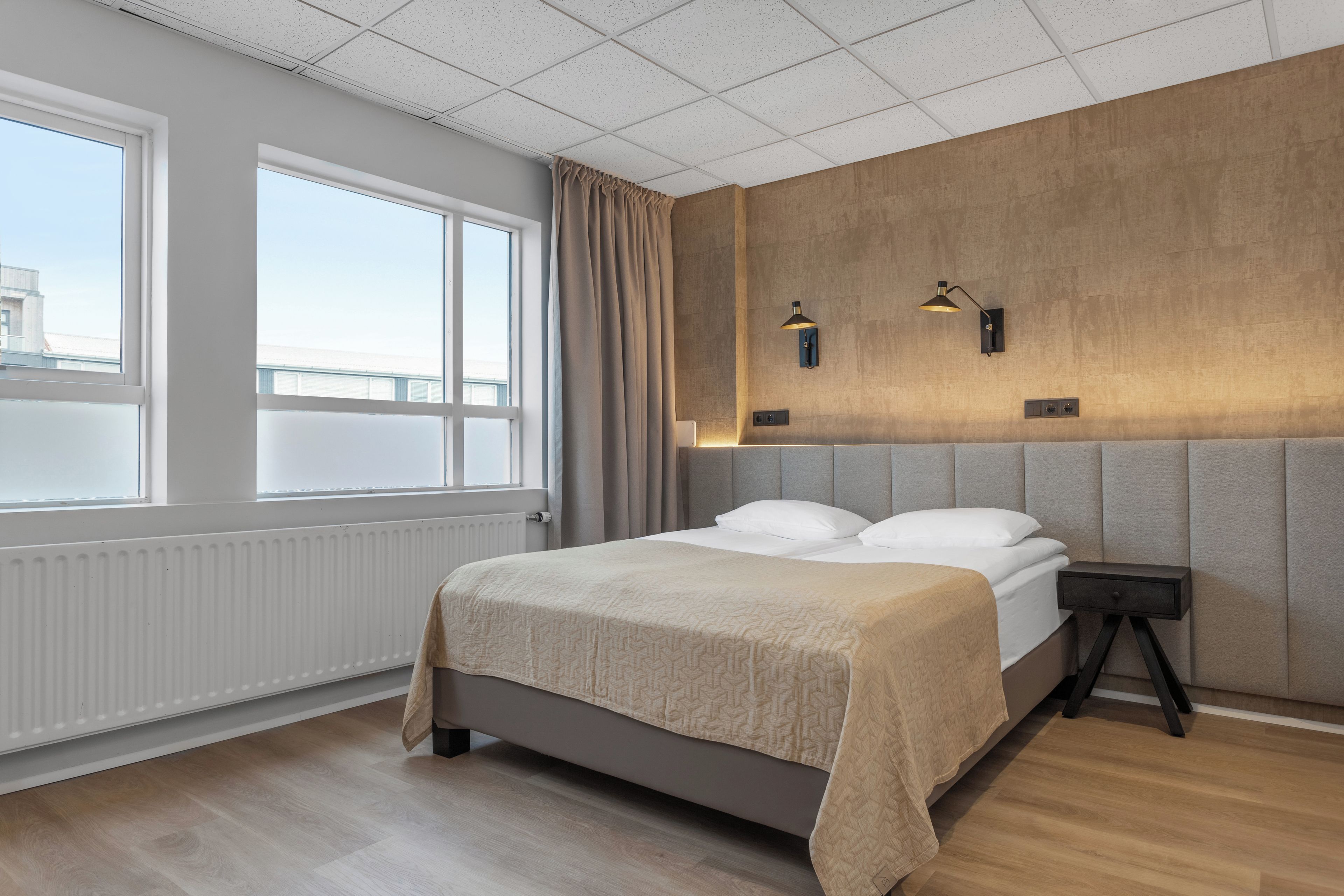 Une chambre moderne avec un grand lit, un radiateur, une fenêtre avec des rideaux et un luminaire fixé au mur.