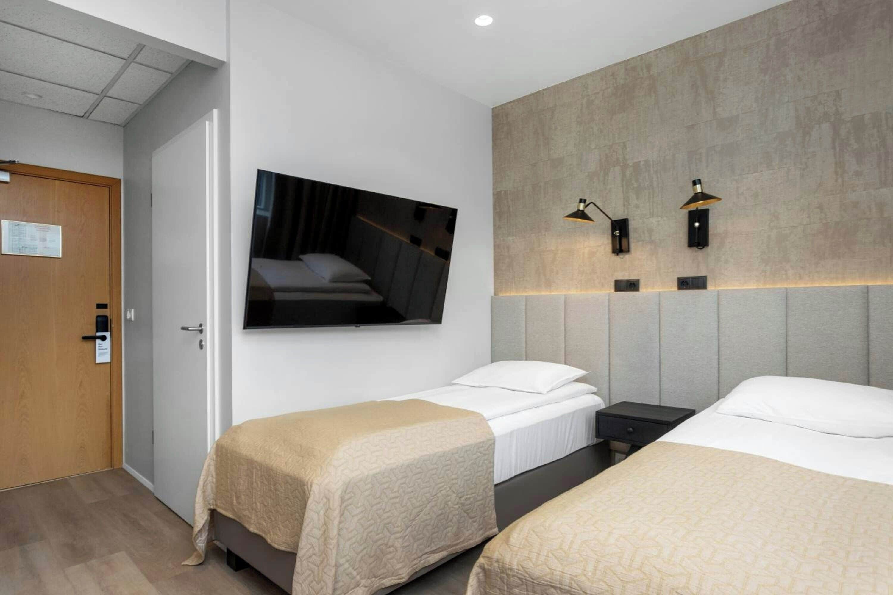 Habitación de hotel con dos camas individuales, un televisor montado en la pared y dos lámparas de pared.