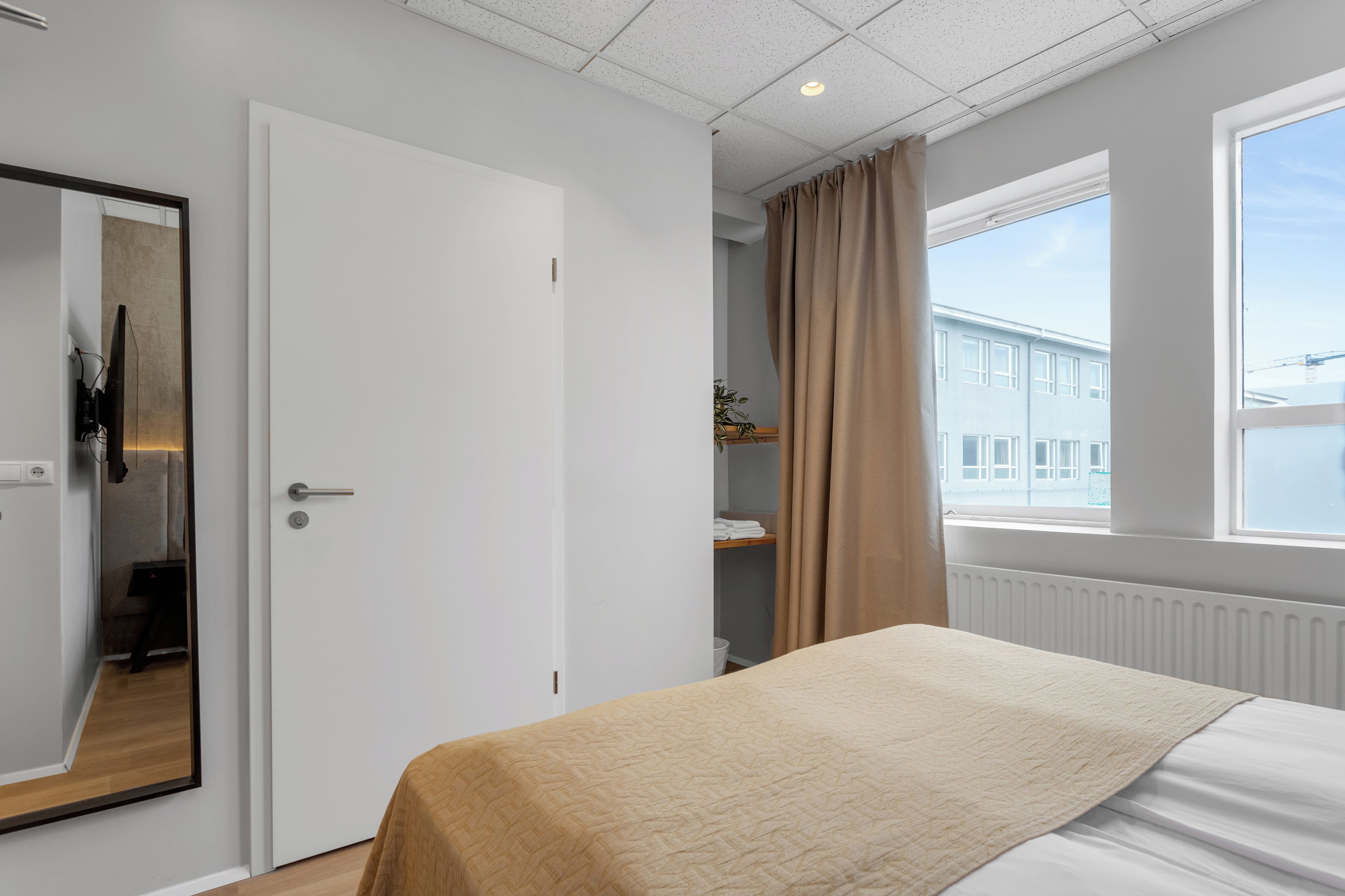 A hotel room with a bed, a door, a window with curtains, and a view of a building outside.