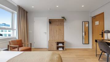 A modern hotel room with a bed, a wooden wardrobe, a desk with a chair, and a large window.