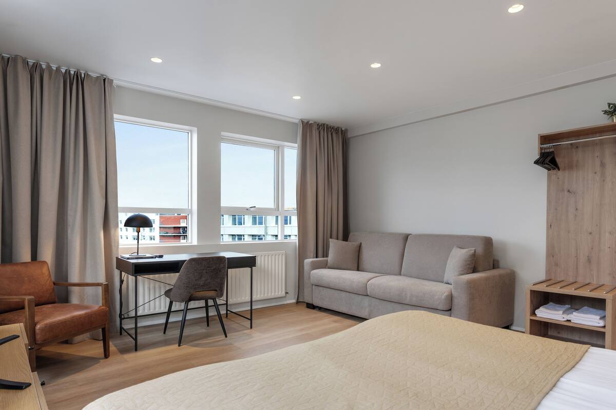 Moderni hotellihuone, jossa on suuri sänky, sohva, työpöytä tuolilla ja suuret ikkunat, joista avautuu näkymä kaupunkiin.