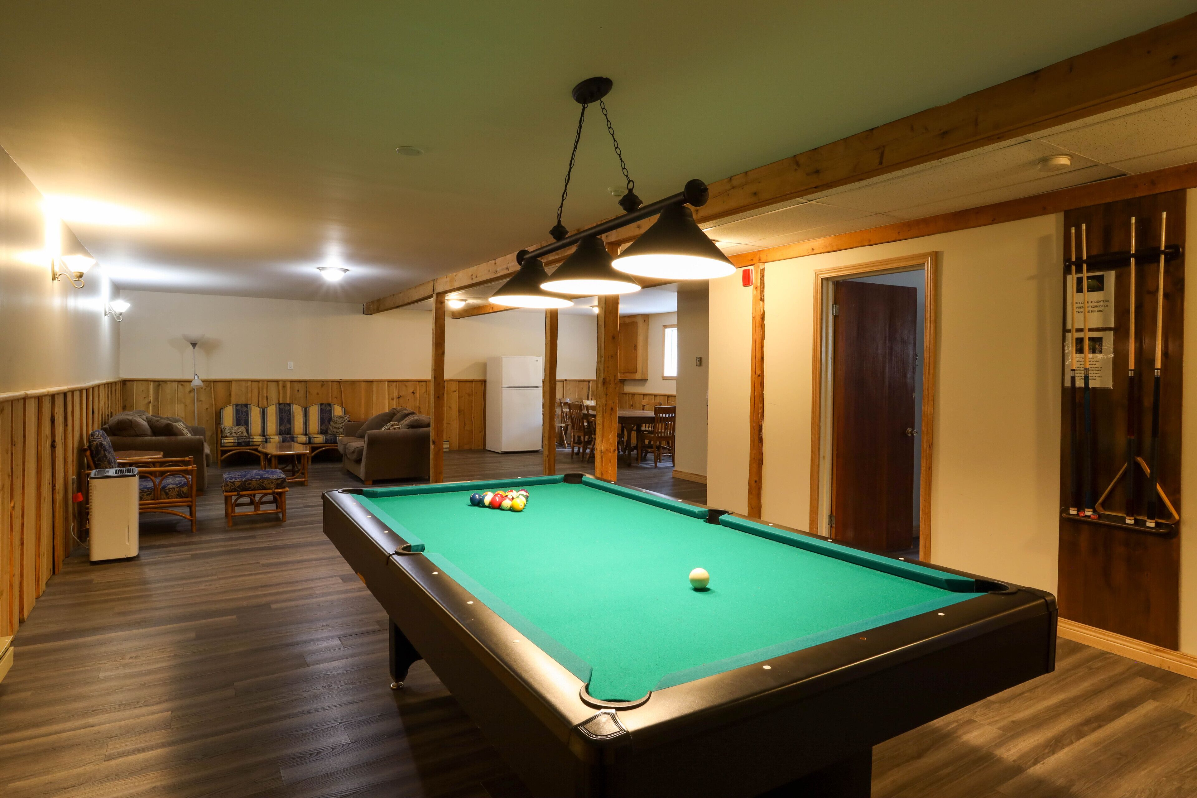 A pool table in a room with wooden walls and ceiling, a seating area with sofas and chairs, and a door leading to another room.