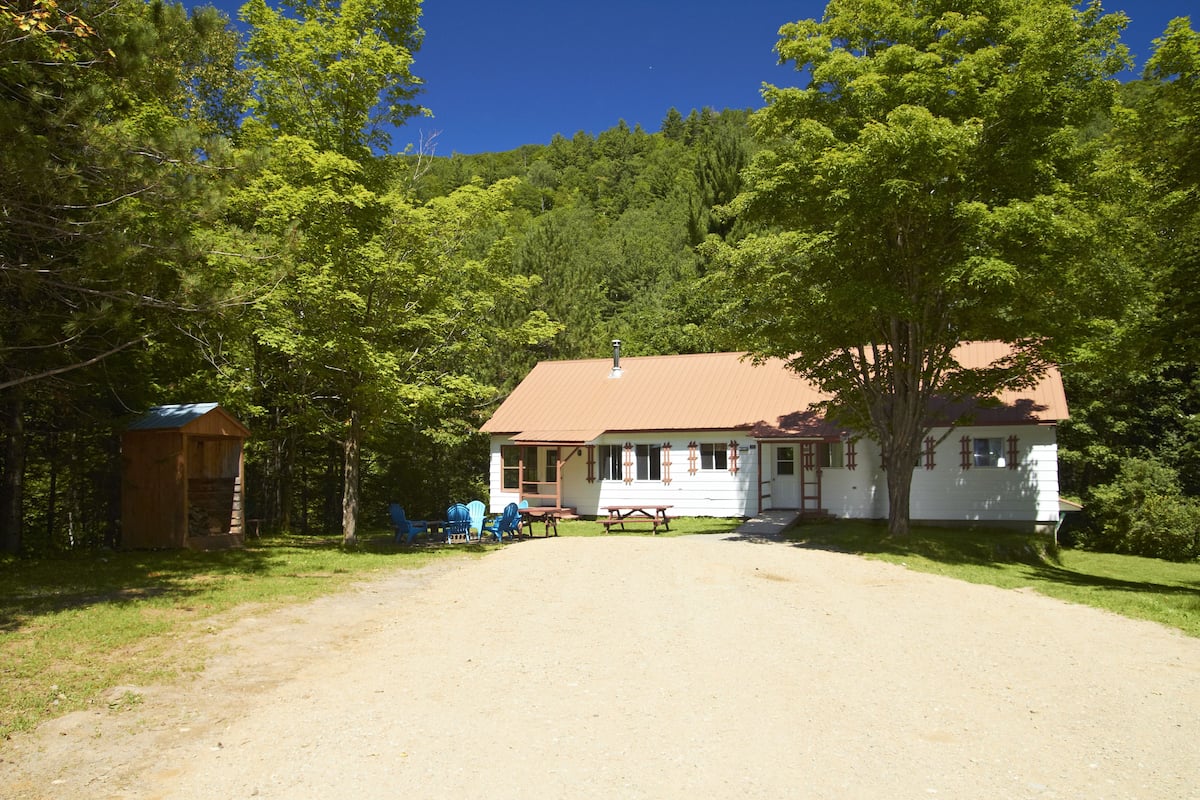 A white building with a brown roof, surrounded by trees. There is a picnic table and blue chairs outside.