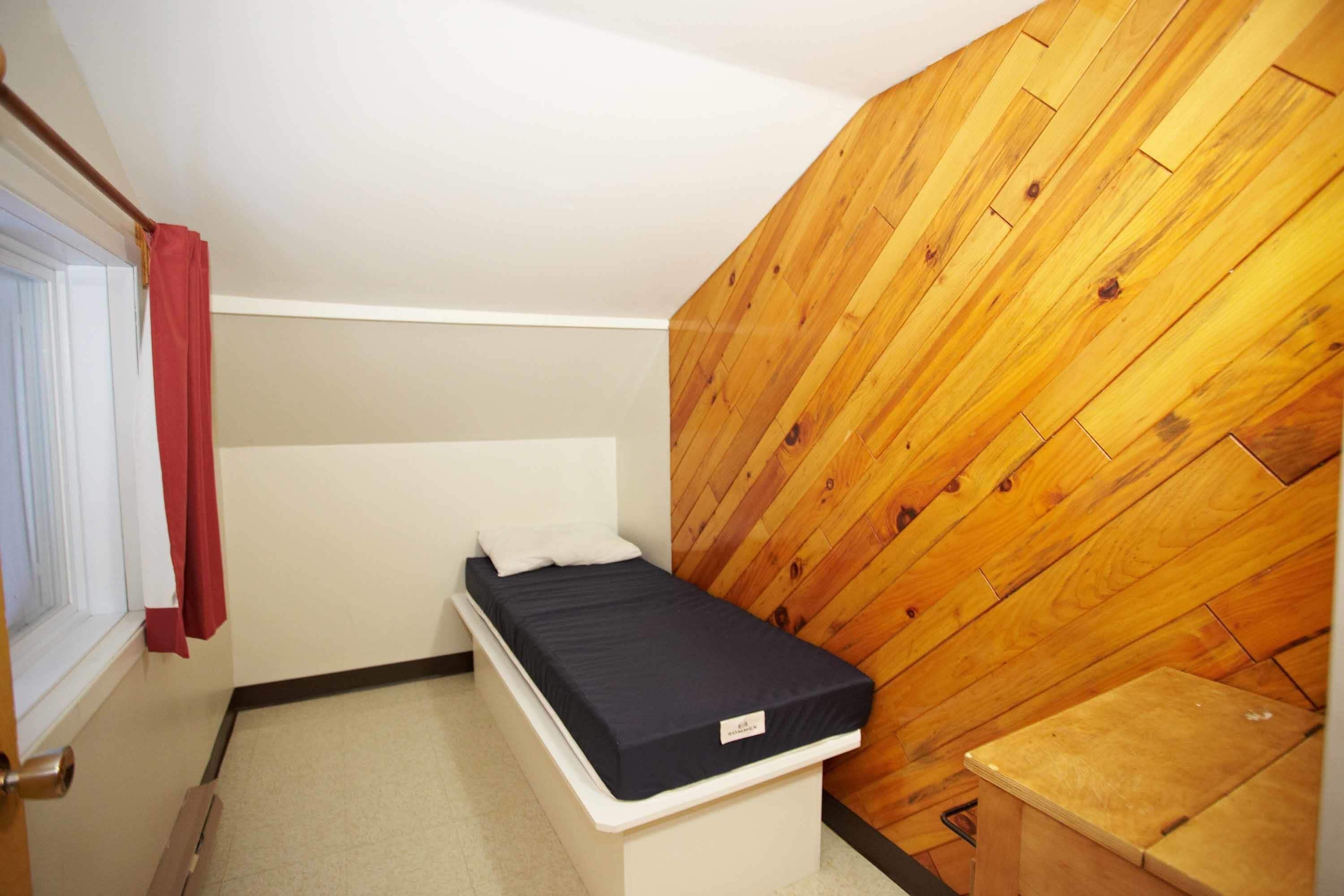 A small, compact room with a single bed, a wooden wall panel, and a small wooden table.