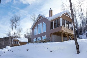 A two-story house with a large deck and a covered porch, surrounded by snow and trees. - Chalets Lanaudiere (Rawdon)