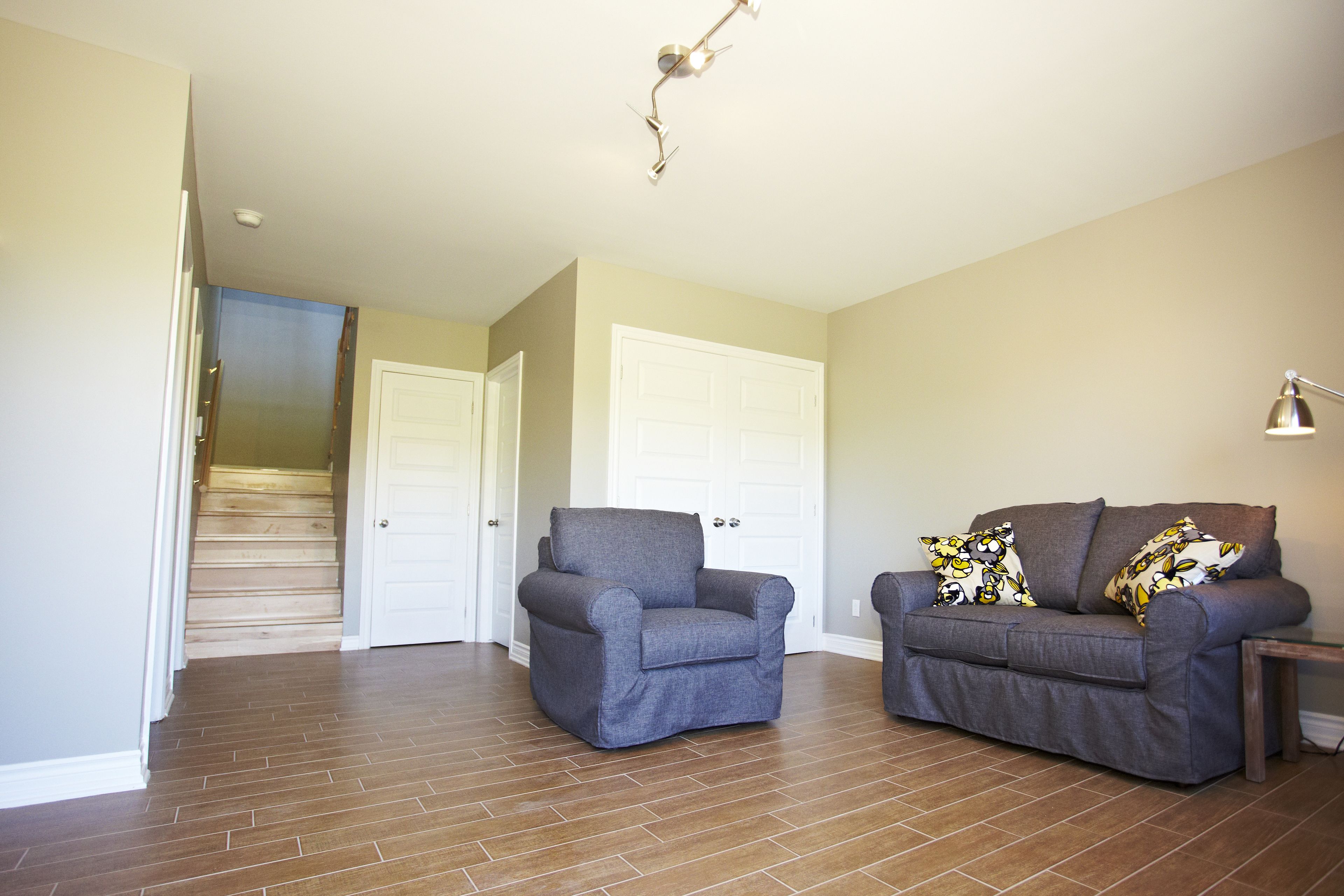 Un salón con un sillón gris y un sofá, una escalera de madera y una puerta blanca al fondo.