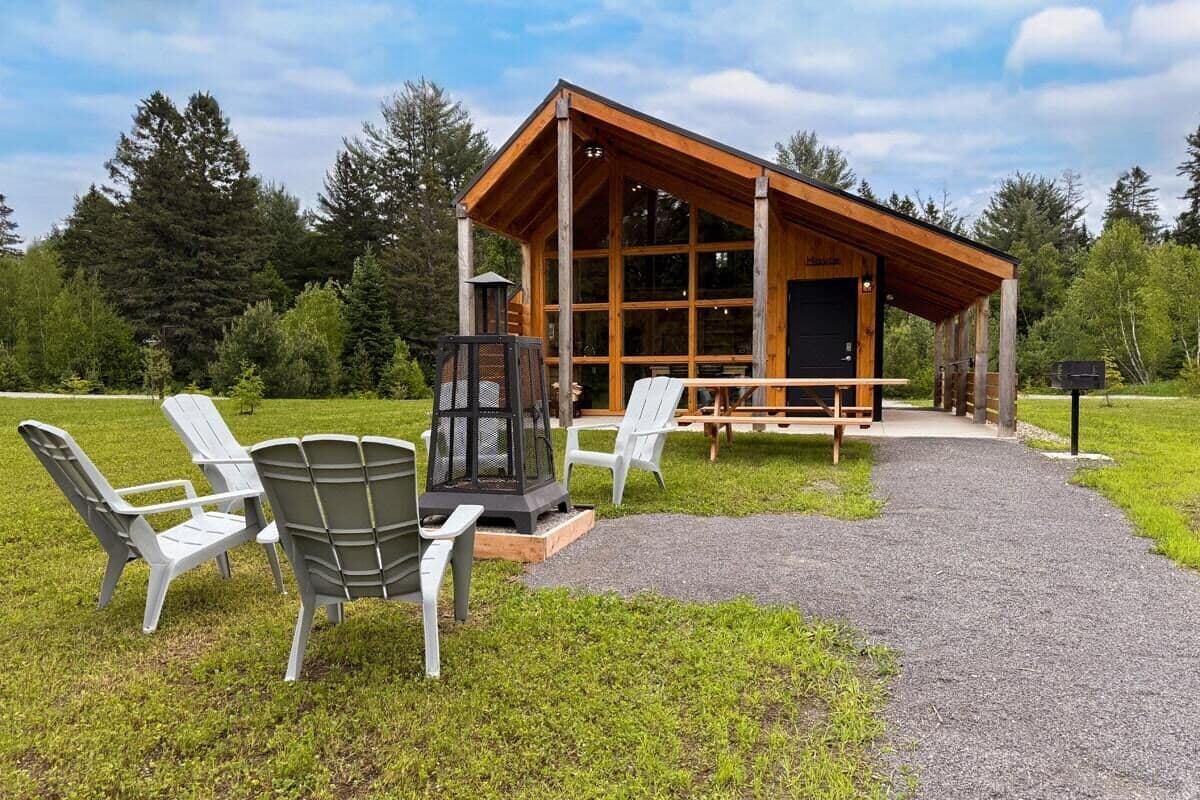 A wooden cabin with a large glass front, surrounded by a gravel path and outdoor seating.
