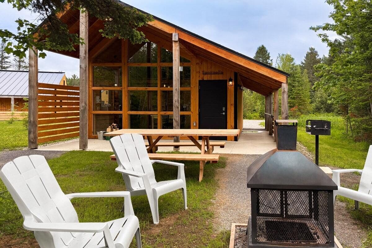 A cabin with a large windowed front, wooden deck, picnic table, and outdoor seating.