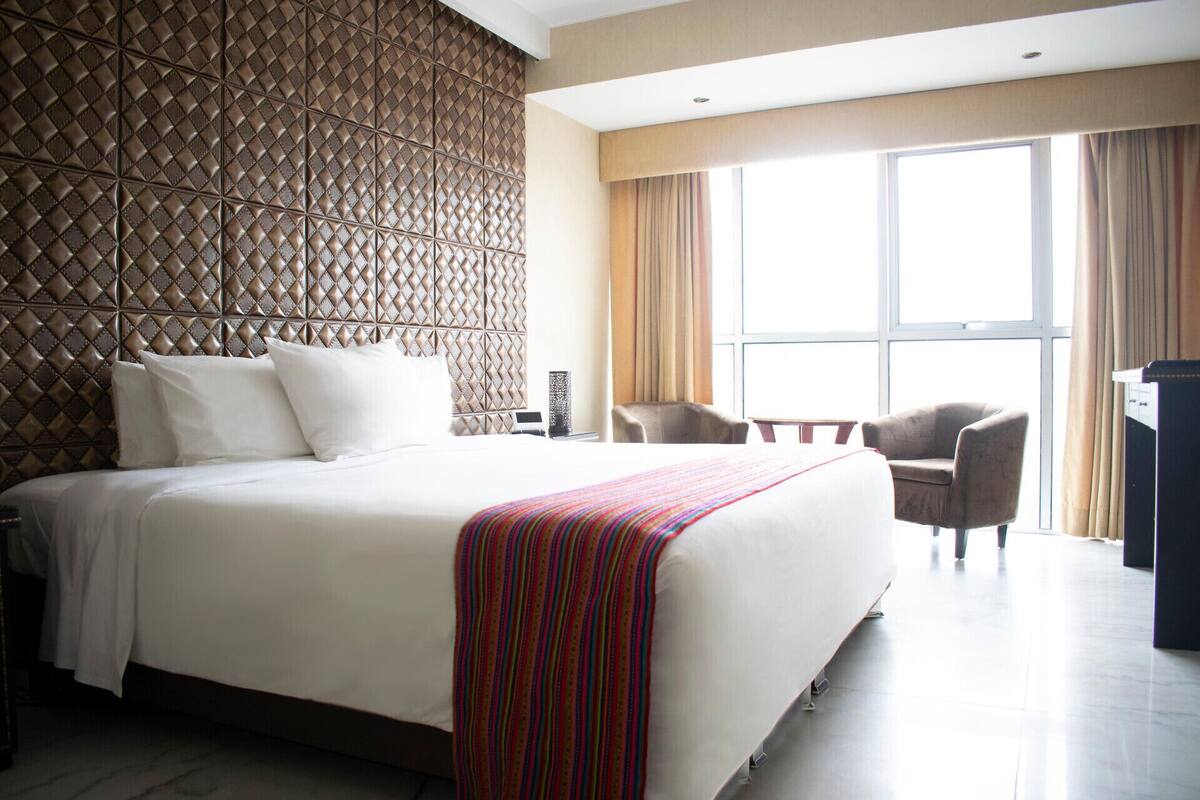 A hotel room with a large bed, a headboard with a diamond pattern, a desk, a chair, and a large window with curtains.