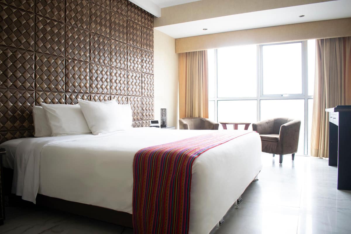 Una habitación de hotel con una cama grande, cabecera con diseño de diamantes, un escritorio, una silla y un ventanal con cortinas.