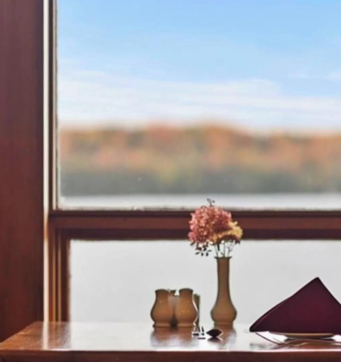 Una ventana con vistas al paisaje, un florero con flores y un objeto en forma de pirámide sobre una mesa.