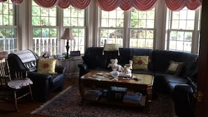 A living room with a dark leather sofa, a wooden coffee table, and a patterned rug. There are large windows with curtains, a lamp, and a decorative horse statue. - The Boston 'T' B&B (Ottawa)