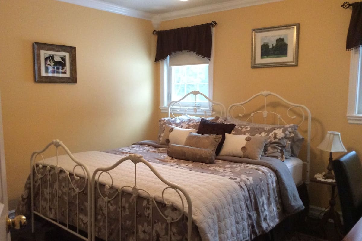 A bedroom with a metal bed frame, floral bedding, a window with curtains, a bedside table with a lamp, and a framed picture on the wall.