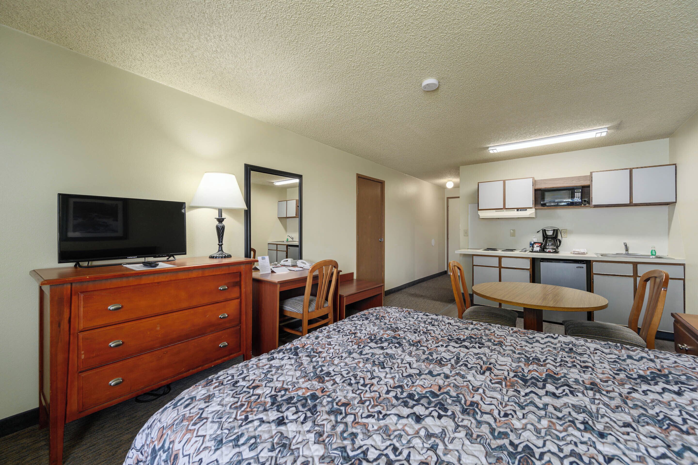 A hotel room with a bed, a desk, a television, a mirror, a kitchenette, and a dining table.