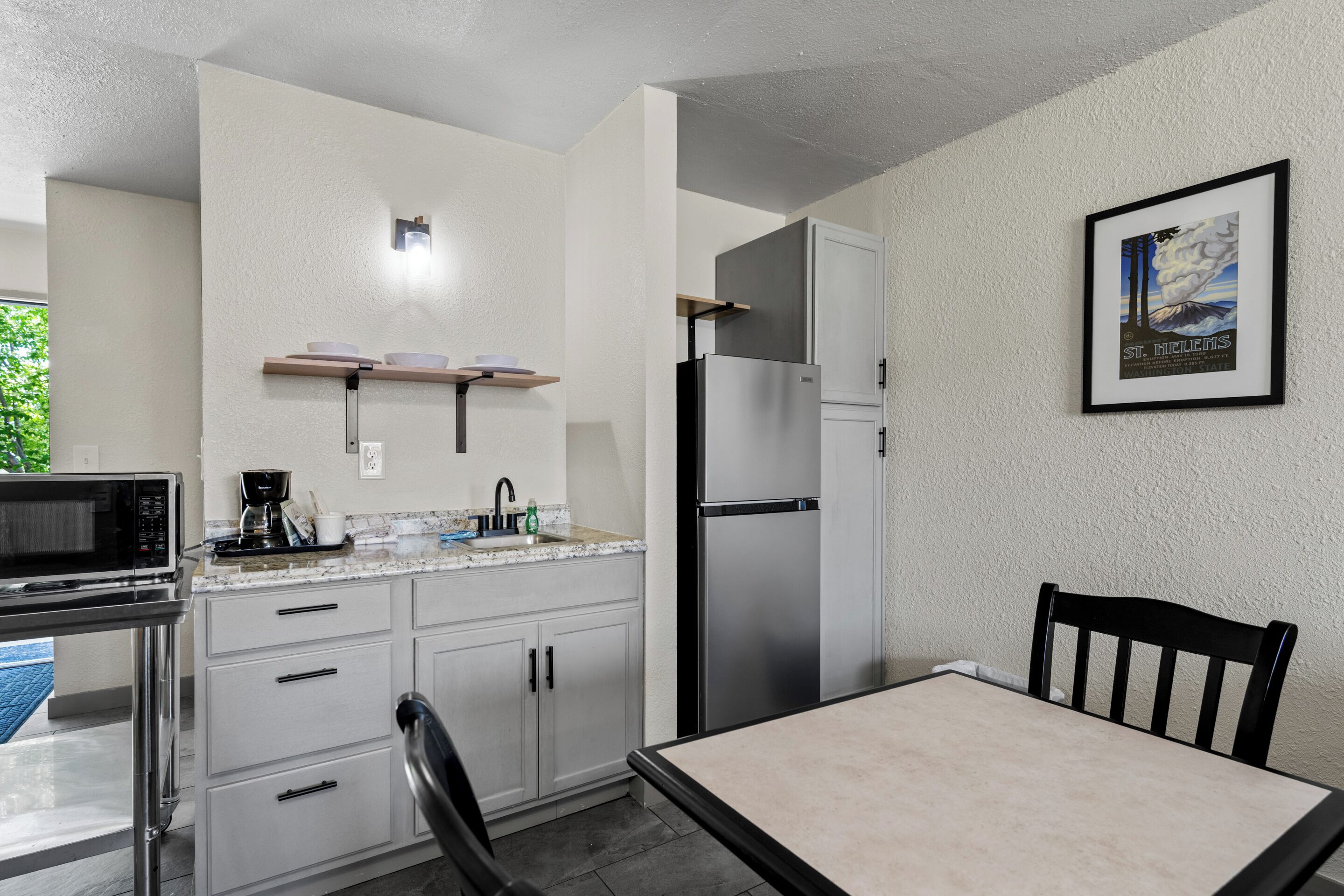 Una cocina compacta equipada con microondas, cafetera y refrigerador. Hay una mesa de comedor con sillas y un cuadro en la pared.