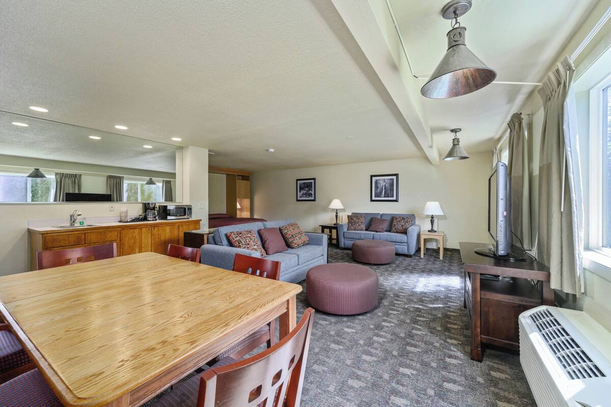A spacious living area with a wooden dining table, a sofa set, and a kitchen area in the background.
