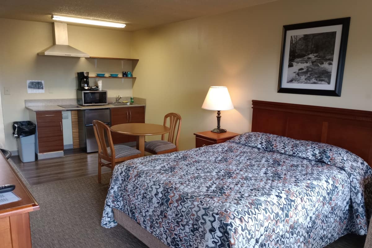 A hotel room with a bed, a desk, a chair, a small kitchenette, and a lamp.
