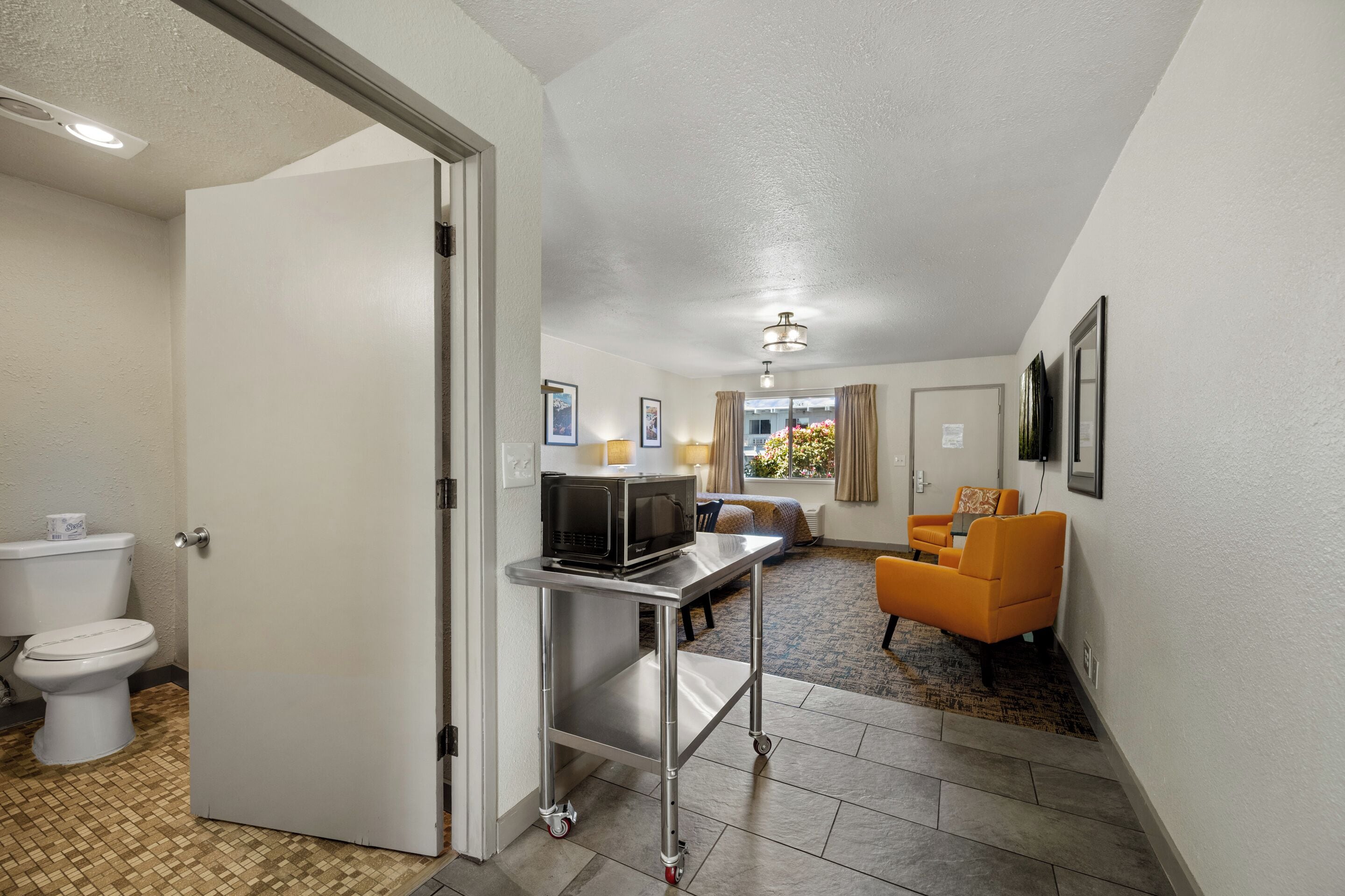 A hotel room with a bathroom, a kitchenette, a seating area with an orange chair, and a window with curtains.