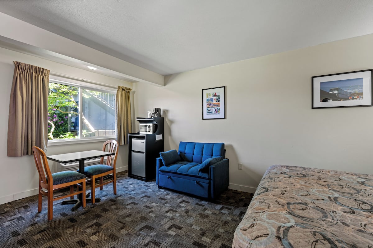 A room with a bed, a desk with a chair, a blue sofa, a coffee machine, and a window with curtains.