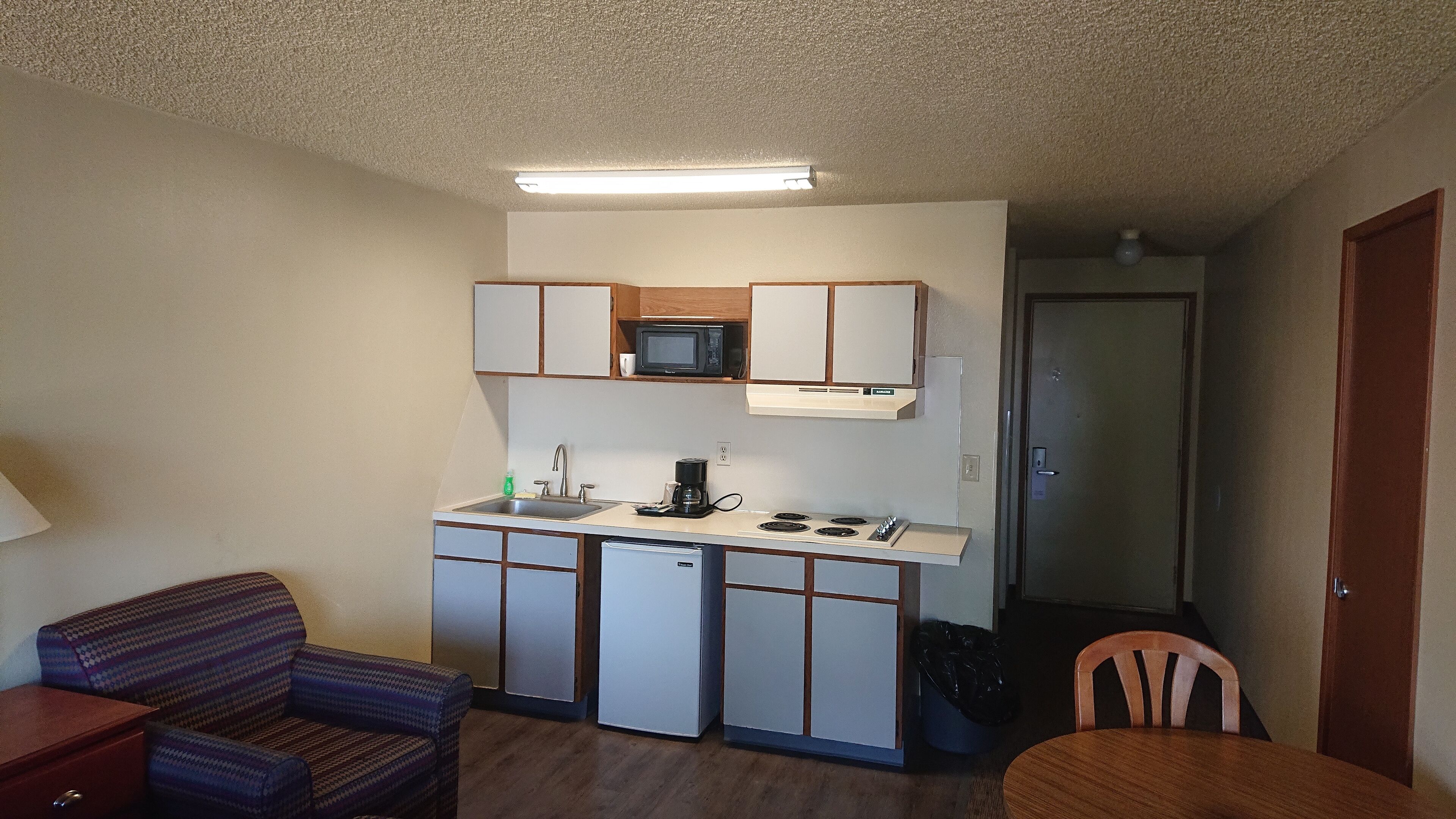Cocina compacta con microondas, pileta y estufa. Incluye un refrigerador pequeño, una silla y una mesa.