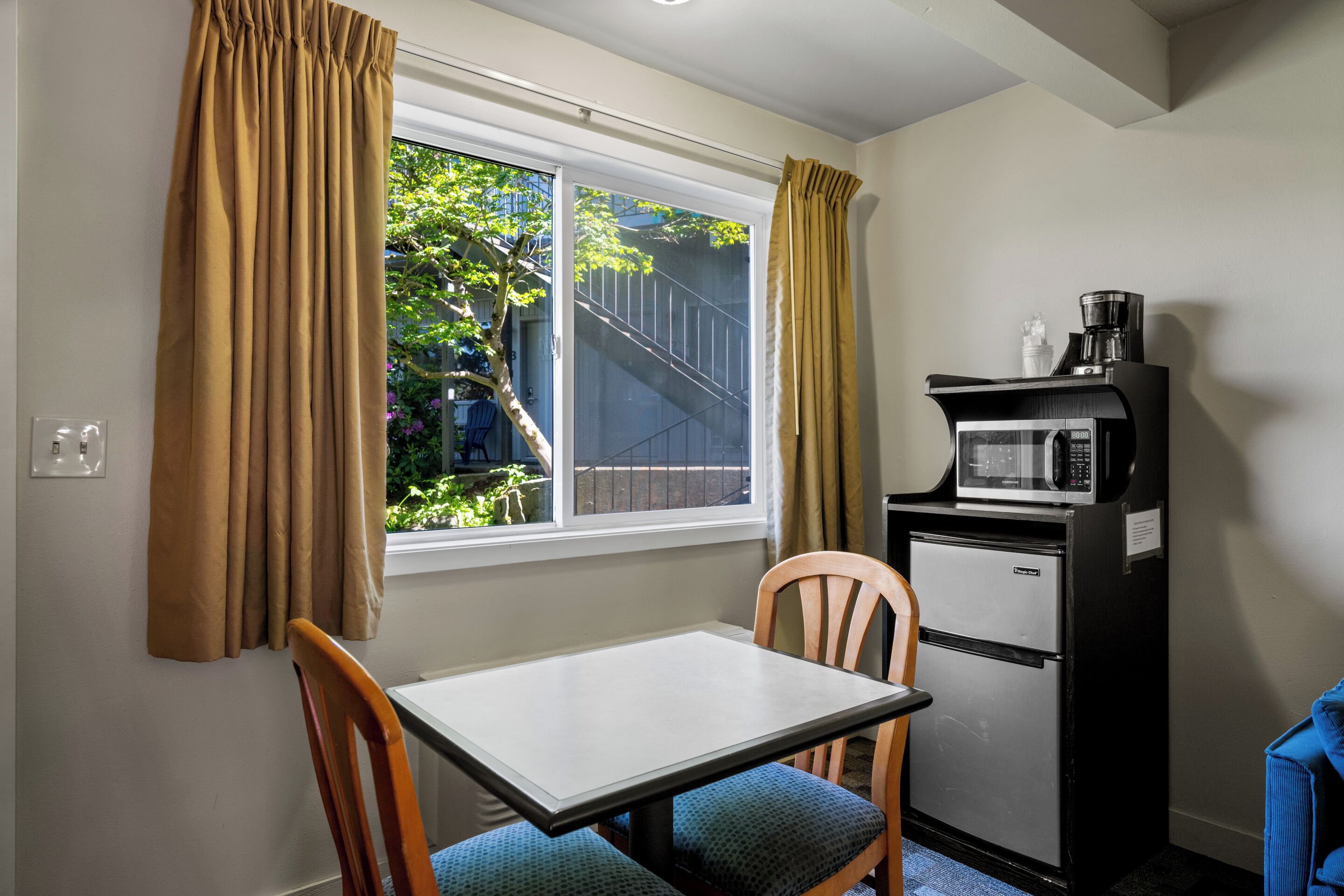 A small kitchenette with a microwave, refrigerator, and coffee maker. There is a window with curtains, a table, and two chairs.