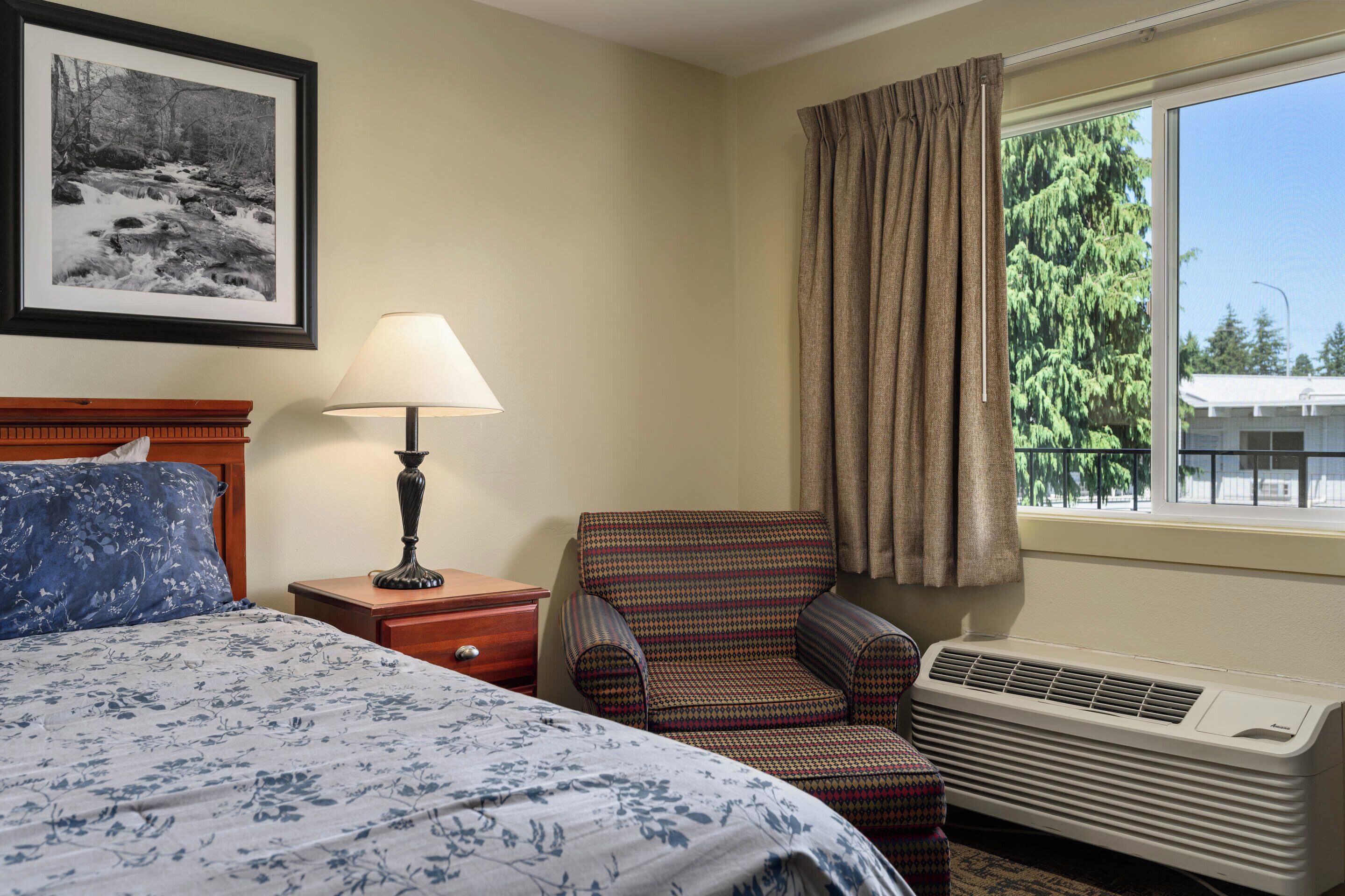 A hotel room with a bed, a chair, a lamp, a heater, a window with curtains, and a framed picture on the wall.