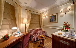 Ein Hotelzimmer mit einem Holztisch, einem gestreiften Sofa, einem kleinen runden Tisch, einer Wandlampe, einem Gemälde und einer Vase mit Blumen.