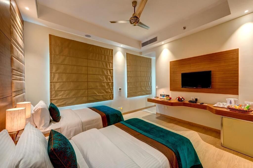 A hotel room with two beds, a TV, a desk with a coffee maker, and large windows with blinds.