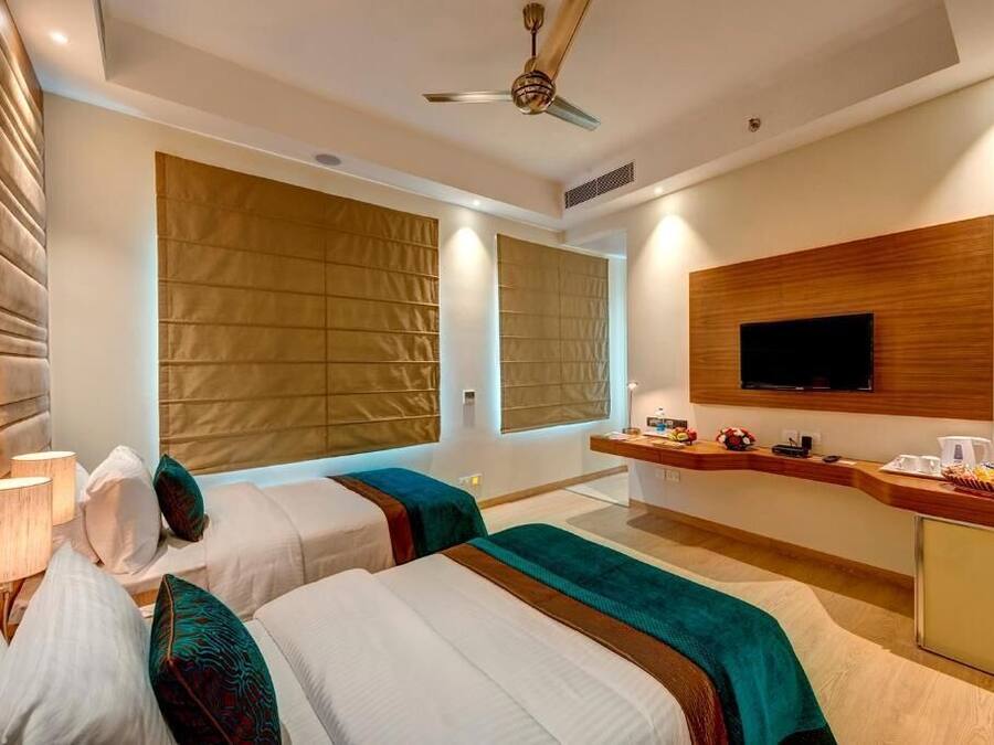 A hotel room with two beds, a TV, a desk with a coffee maker, and large windows with blinds.