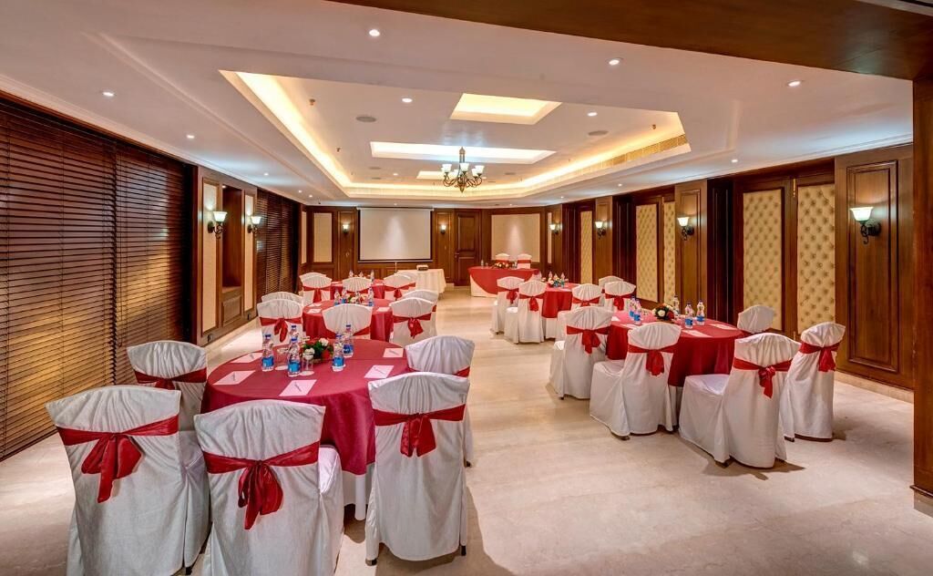 Un salón de conferencias con mesas redondas, sillas blancas con cintas rojas y una pantalla de proyector.