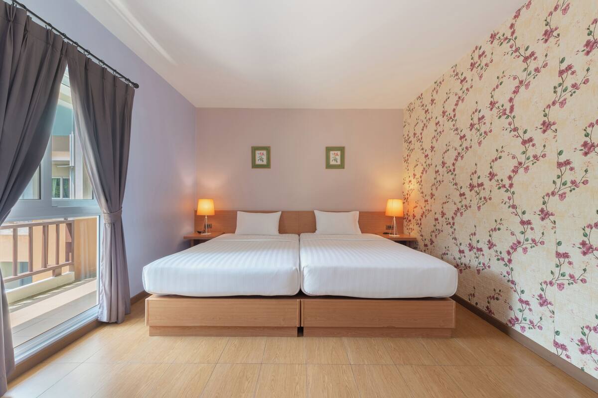 A double bed with wooden headboard, two bedside lamps, floral wallpaper, and a balcony view.