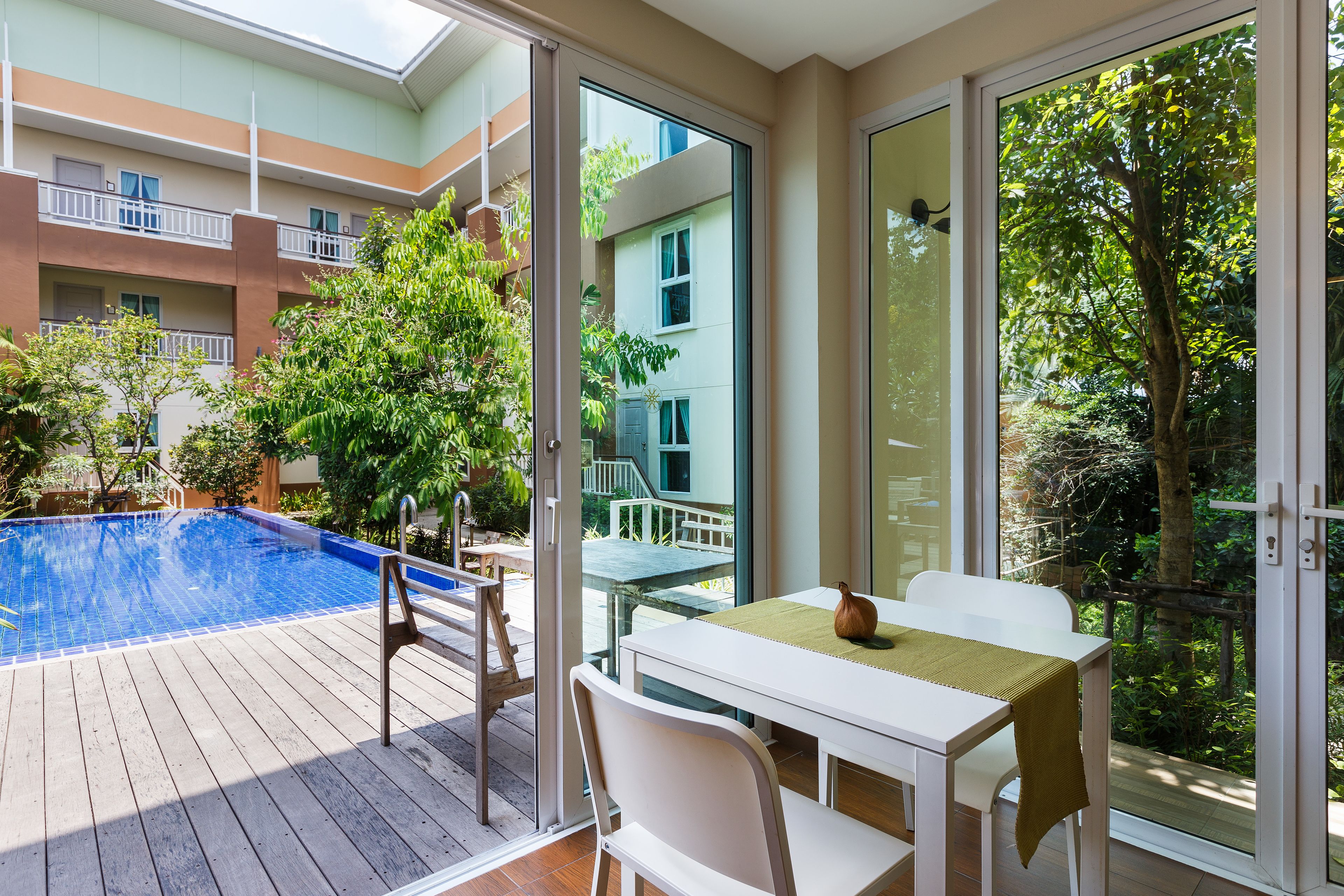 A poolside area with a table and chairs, a view of a building with balconies, and a tree in the background.