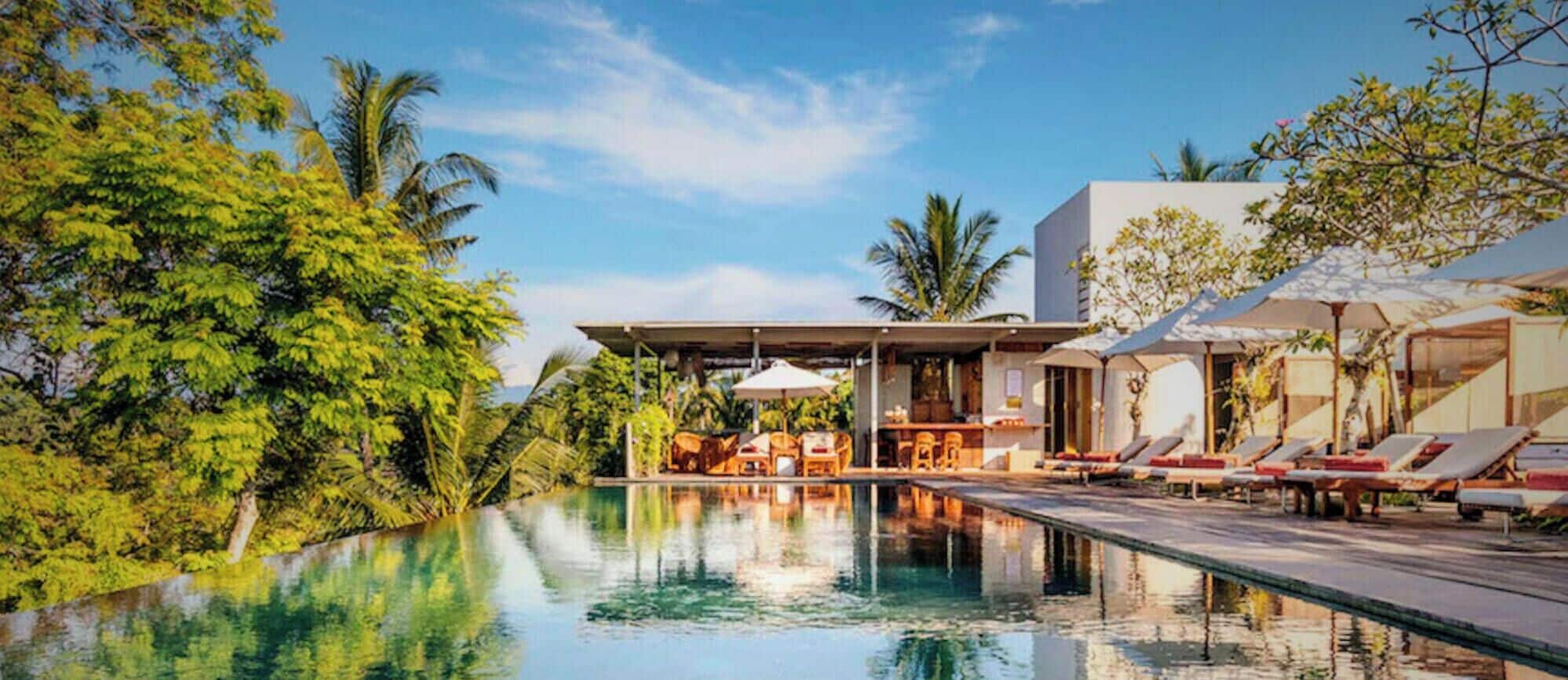 A pool area with lounge chairs and umbrellas, surrounded by lush greenery and a modern building structure.