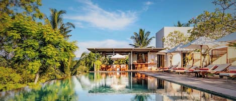 Un área de piscina con sillones de descanso y sombrillas, rodeada de abundante vegetación y una estructura de edificio moderna.