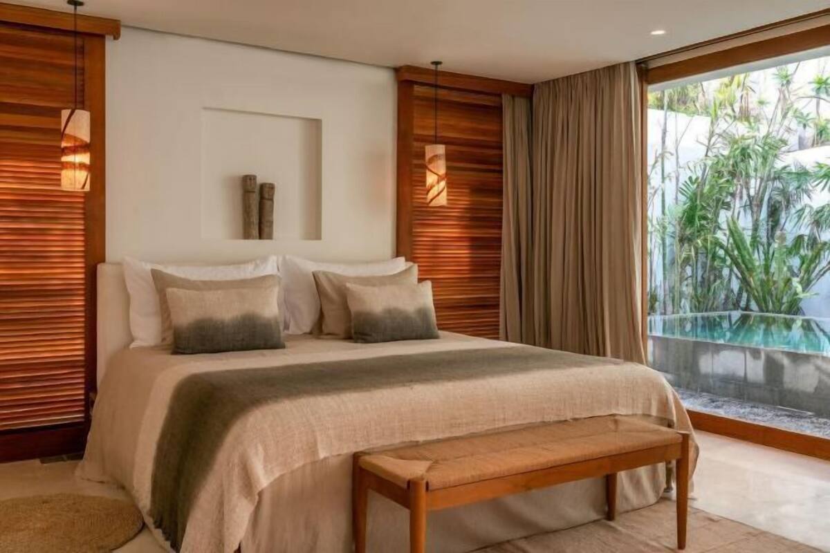 A bedroom with a large bed, a bench, wooden paneling, and a view of greenery through a glass door.