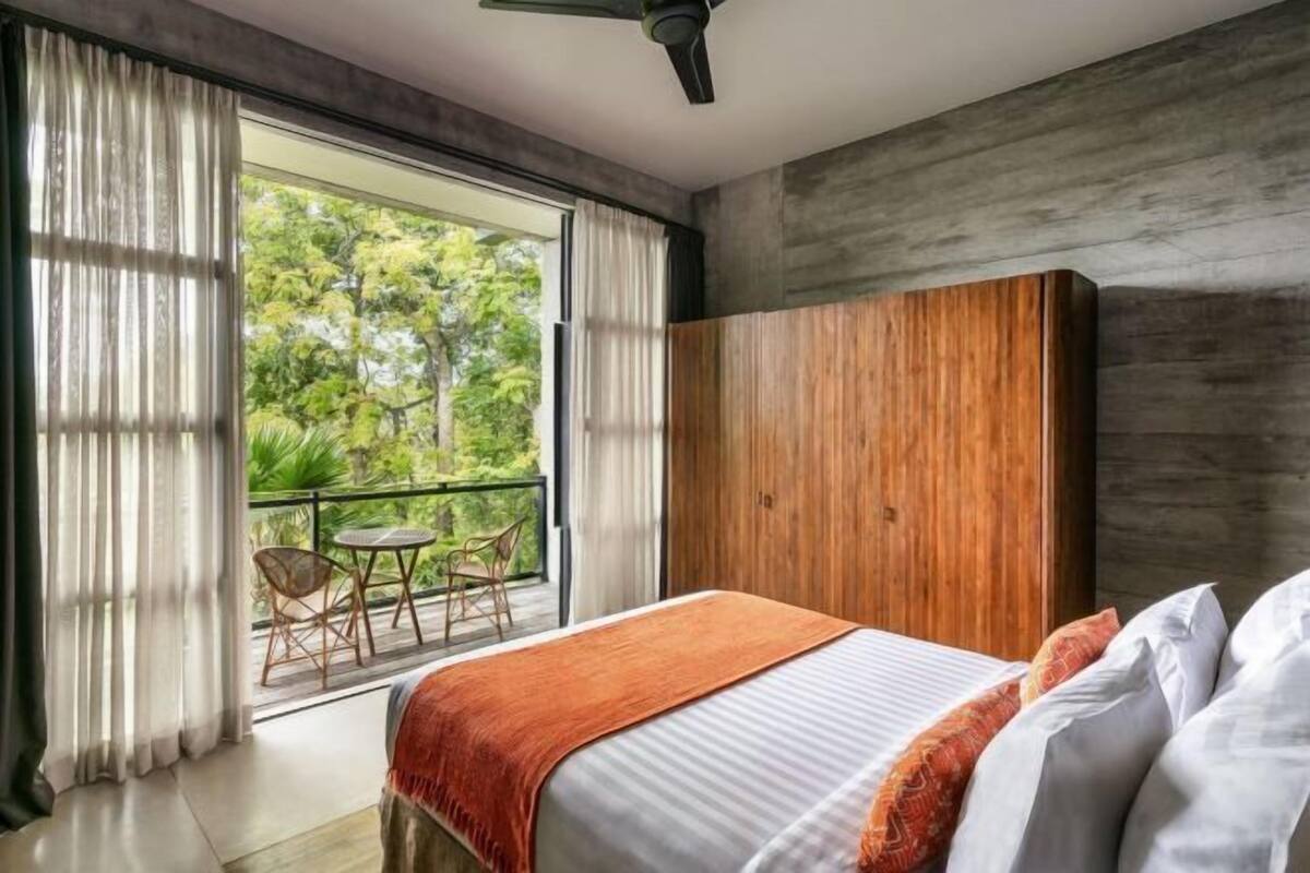 A bedroom with a bed, a wooden wardrobe, a balcony with chairs, and a view of greenery.