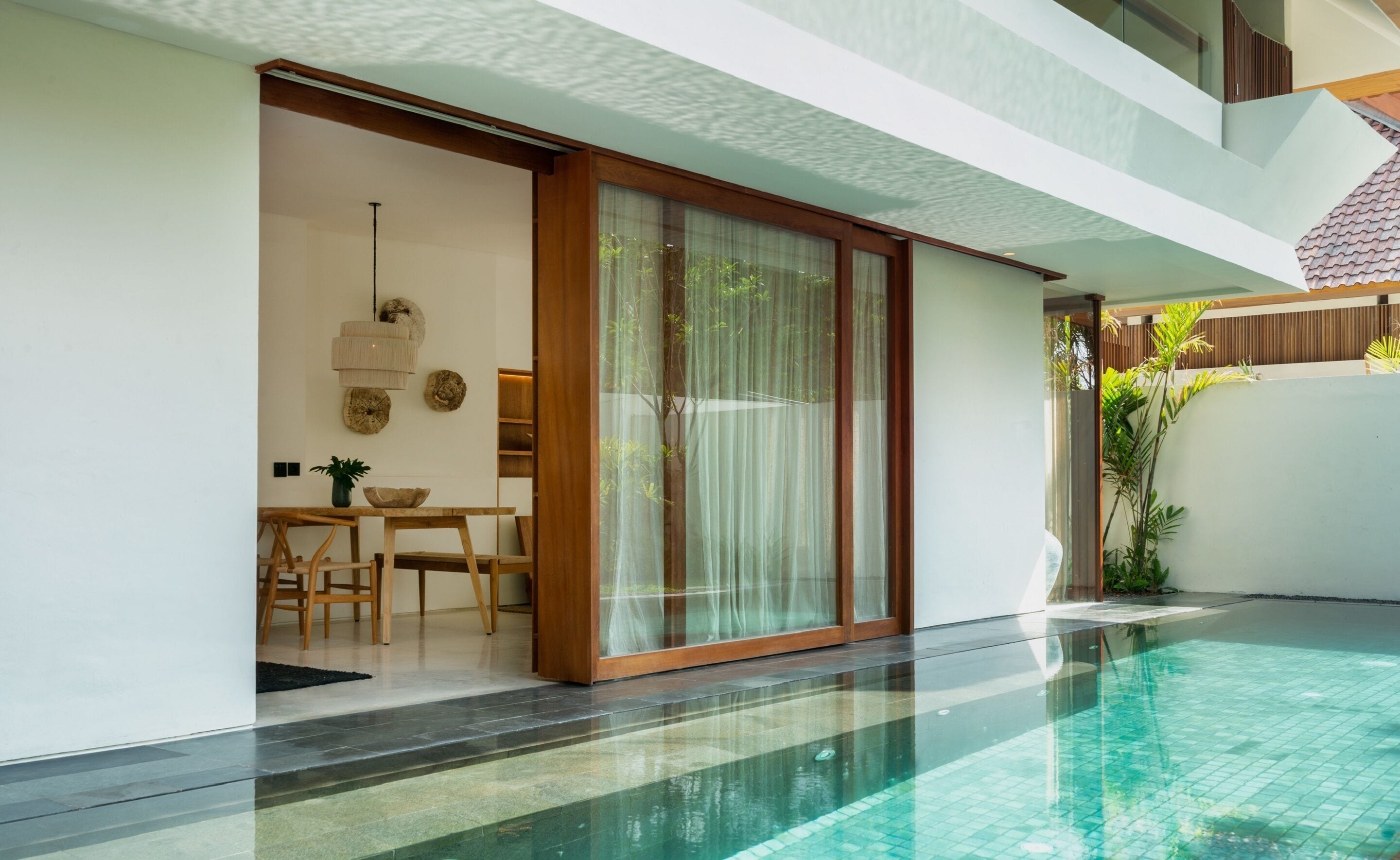 A modern poolside area with a glass-enclosed dining space, a wooden table, and a clear pool.