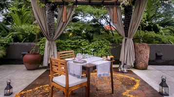 An outdoor dining area with a table set for two, surrounded by greenery and a pergola.